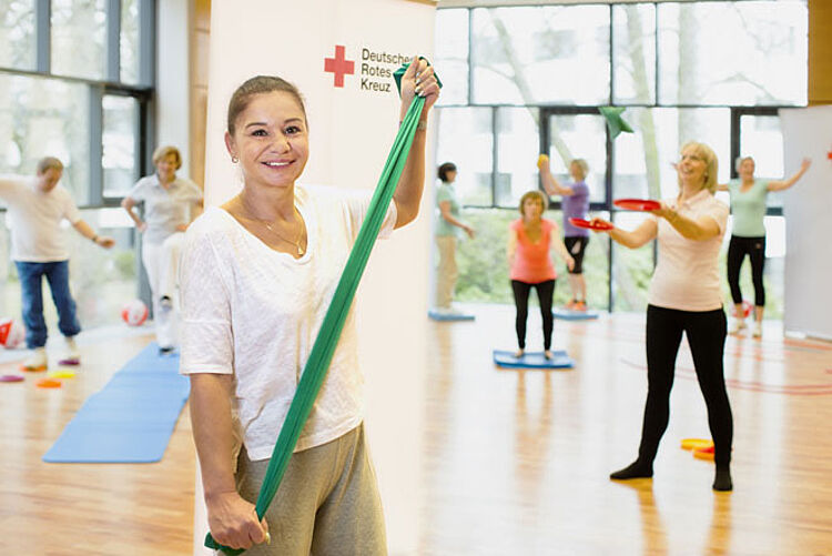 Gesundheitskurs, Gymnastik Kurs Eine Kursleiterin hält ein elastisches Band lächelnd in die Kamera.